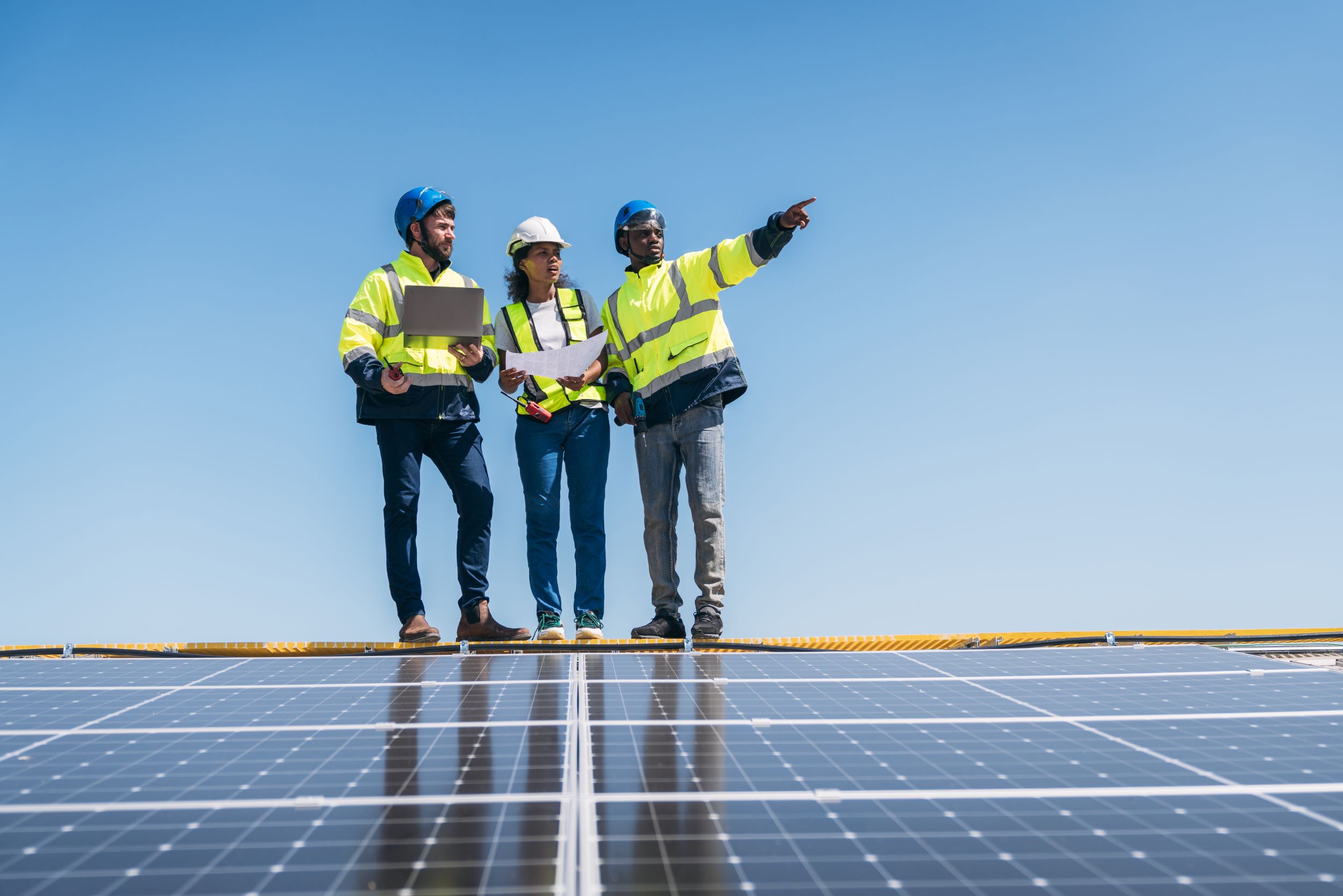 3 mensen die bij zonnepanelen staan, met papieren en een laptop waarvan 1 wijst naar iets